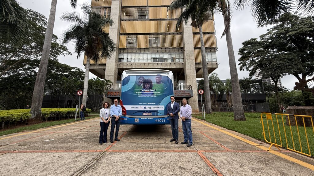 Foto em plano mais fechado mostrando o grupo em frente à traseira do ônibus da campanha Declare o Bem. O ônibus tem o fundo azul com rostos sorridentes de três pessoas representando diversidade. A frase central diz: “Seu imposto de renda pode transformar vidas”. Da esquerda para a direita, estão: Cris Martins (gerente do Fundo Social), o prefeito Helinho Zanatta, Evandro Amaral (presidente do Conselho de Administração da Sicoob Cocre) e Odair Mello (secretário de Segurança Pública, Trânsito e Transportes), todos em trajes sociais.