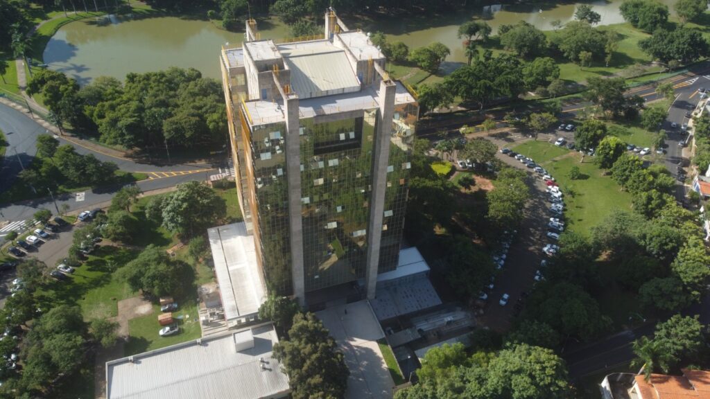 A imagem mostra uma vista aérea de um edifício alto cercado por áreas verdes, estradas e um estacionamento. O edifício tem janelas de vidro reflexivas e um telhado plano com algumas estruturas no topo. Ao fundo, há um grande lago ou reservatório, rodeado por árvores e gramados. As estradas próximas ao prédio têm carros estacionados, e há vários veículos no estacionamento adjacente ao edifício. A