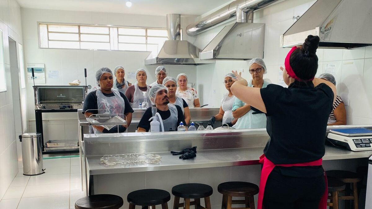 Instrutora fala para uma turma de alunos reunidos em uma cozinha industrial. Os participantes usam toucas higiênicas e aventais, atentos à explicação em frente ao fogão e bancadas.