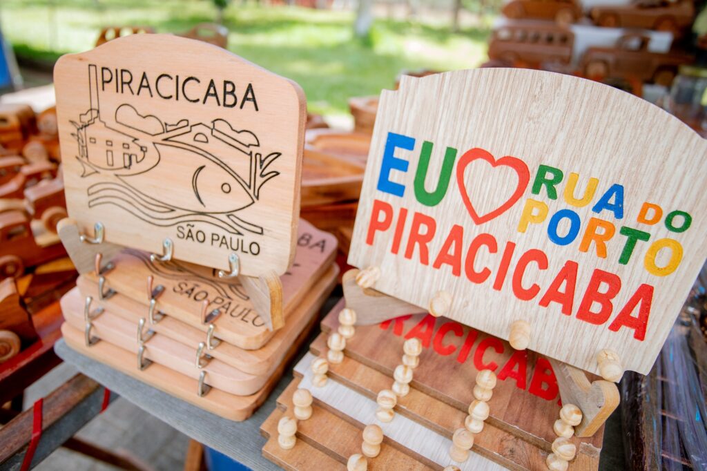 A imagem mostra uma variedade de artesanato de madeira à venda, provavelmente em uma feira ou mercado. O foco principal está em dois tipos de placas de madeira:* *Placas com imagem e texto:* Um conjunto de placas de madeira empilhadas apresenta uma imagem gravada de um peixe e uma cidade ribeirinha, com "PIRACICABA" e "SÃO PAULO" escritos na placa. As placas têm ganchos, sugerindo que podem ser usadas como porta-chaves ou organizadores.* *Placas com texto colorido:* Uma placa de madeira separada tem a frase "EU ❤️ RUA DO PORTO PIRACICABA" escrita em letras coloridas, com o coração vermelho como o elemento central. A palavra "PIRACICABA" é a maior e mais proeminente. Esta placa está em um suporte, e parece ser uma peça única, ou pelo menos não empilhada como as outras.Além dessas placas, há outras peças de artesanato de madeira ao redor, incluindo o que parecem ser pequenas peças de madeira em forma de contas ou botões, e outras placas de madeira de tamanhos e formas variadas, algumas das quais estão ligeiramente desfocadas. O fundo sugere um ambiente externo, com um gramado verde borrado ao fundo, indicando que o mercado pode estar em um espaço aberto. A iluminação parece natural, sugerindo luz do dia.A imagem sugere que os artesanatos são souvenirs ou itens decorativos relacionados à cidade de Piracicaba, São Paulo, Brasil. A variedade de peças e o agrupamento sugerem uma seleção de produtos disponíveis para compra.