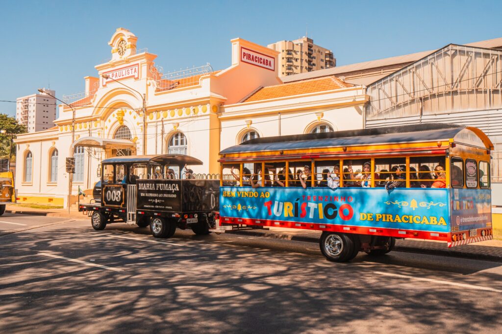 A imagem mostra um trem turístico chamado "Trenzinho Turístico de Piracicaba," estacionado em frente a um edifício histórico identificado como "Estação da Paulista". O trem tem uma locomotiva preta com o nome "Maria Fumaça" e o número "003," seguida por um vagão de passageiros colorido com a frase "Bem Vindo ao Trenzinho Turístico de Piracicaba." O edifício ao fundo possui estilo arquitetônico clássico, com janelas em arco e elementos decorativos. O cenário está iluminado pelo sol, com outros edifícios visíveis ao fundo.