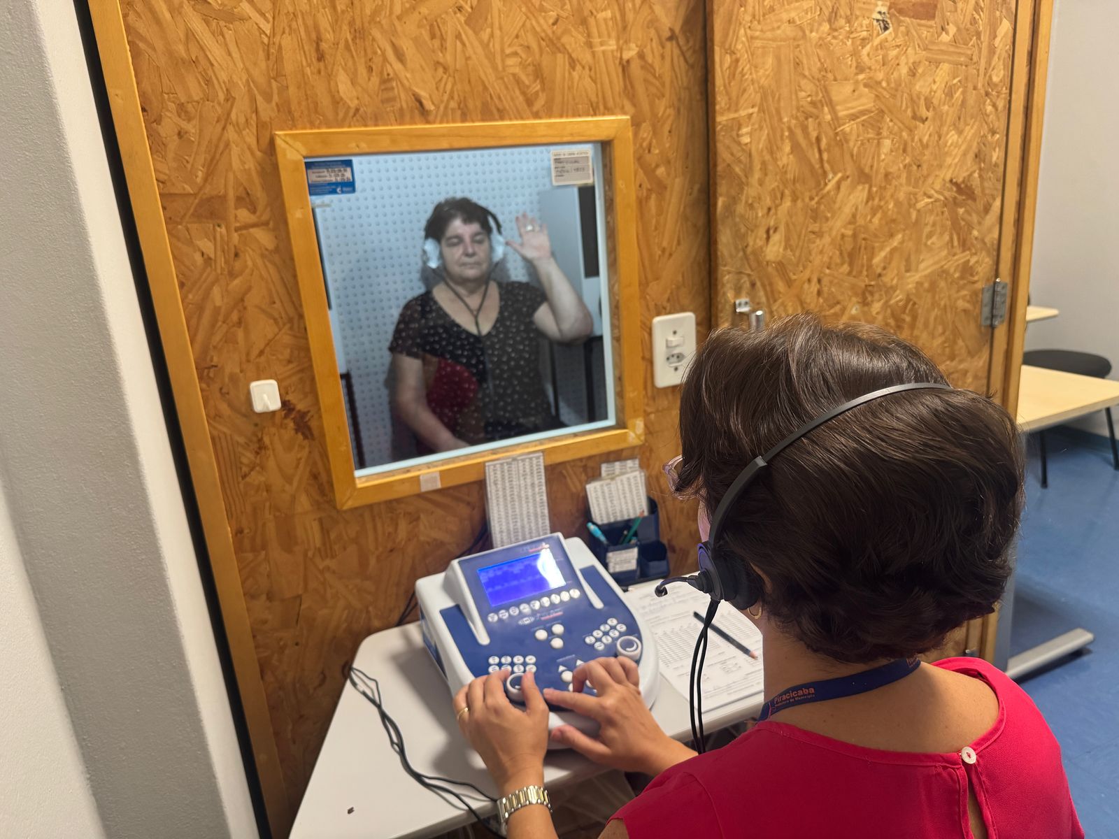 A imagem mostra um exame de audiometria. Vamos analisar os detalhes: *O que está acontecendo?* Uma audiometrista (a pessoa de costas, usando fones de ouvido e operando um equipamento) está realizando um teste de audiometria em uma paciente (a pessoa visível através da janela insonorizada). A paciente está usando fones de ouvido e sinaliza com a mão quando ouve um som. *Detalhes do processo:* * *A Cabine Inesonorizada:* A paciente está sentada em uma cabine insonorizada, projetada para minimizar ruídos externos que possam interferir no teste. A parede da cabine é geralmente feita de material absorvente de som. A janela permite que a audiometrista observe a paciente. * *O Equipamento:* A audiometrista utiliza um audiômetro, um dispositivo eletrônico que gera sons em diferentes frequências e intensidades. O audiômetro mede a capacidade da paciente de ouvir esses sons. A tela mostra os resultados do teste em tempo real ou dados relevantes para a condução do exame. * *O Teste:* O teste de audiometria avalia a capacidade auditiva da paciente em diferentes frequências. A paciente sinaliza quando ouve um som, permitindo que a audiometrista registre o nível de audição em cada frequência. A resposta manual indica se o som foi percebido ou não. * *A Profissional:* A audiometrista é uma profissional de saúde treinada para realizar exames de audiometria e interpretar os resultados. A credencial visível em sua roupa sugere uma afiliação profissional, provavelmente a uma clínica ou instituição. *Em resumo:* A imagem documenta um exame de audiometria, um procedimento comum para avaliar a saúde auditiva. O processo envolve uma cabine insonorizada, um audiômetro, a participação ativa da paciente e a perícia da audiometrista.