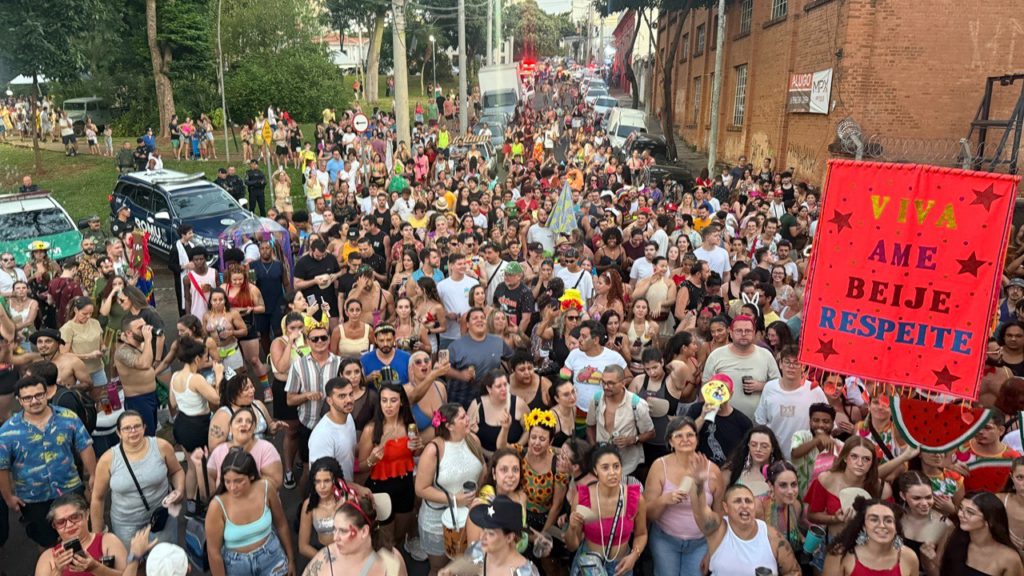 A imagem mostra uma grande multidão de pessoas participando de um desfile de rua ou carnaval. A multidão é densa e composta por indivíduos de diversas idades e vestimentas, muitos usando fantasias e adereços carnavalescos. Há uma atmosfera festiva e animada, com pessoas interagindo umas com as outras.No centro da imagem, um grande banner vermelho chama a atenção, com a mensagem "Viva, Ame, Beije, Respeite" escrita em letras grandes. A mensagem promove a inclusão, o amor e o respeito, valores frequentemente associados a eventos festivos e desfiles de orgulho LGBTQIA+.Há também a presença de veículos policiais, sugerindo uma medida de segurança para o evento. A multidão está se movendo em uma rua, entre prédios, e a vegetação ao fundo sugere que o evento está ocorrendo em uma área urbana com árvores e espaços verdes próximos.Em resumo, a imagem retrata um momento vibrante e inclusivo de um evento público, provavelmente um desfile de rua ou carnaval, com ênfase na mensagem de amor e respeito. A presença policial indica uma preocupação com a segurança e o bom andamento do evento.