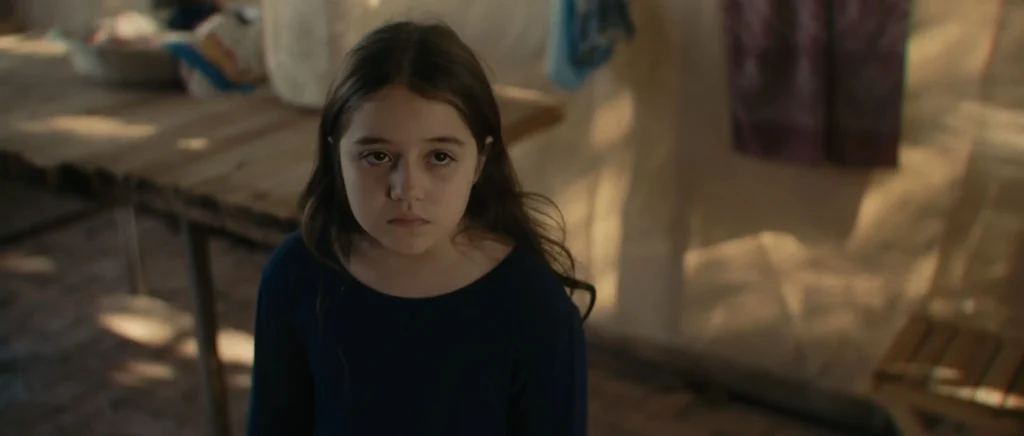 Menina de cabelos longos e escuros olha séria para frente, dentro de um ambiente simples com móveis rústicos.