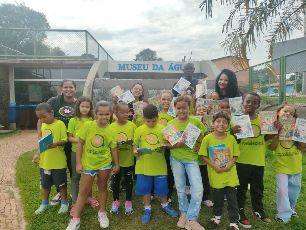 A imagem mostra um grupo de crianças e adultos em frente ao Museu da Água. As crianças estão vestindo camisetas verdes com um logo e segurando livros ou revistas infantis intitulados "Lamba". Há várias pessoas adultas sorrindo, algumas segurando também exemplares da mesma publicação. O cenário indica um passeio ou atividade educativa relacionada à leitura e ao museu. A atmosfera é alegre e descontraída, sugerindo um evento positivo e inclusivo. A imagem captura um momento de interação entre as crianças, os adultos e o ambiente do museu.