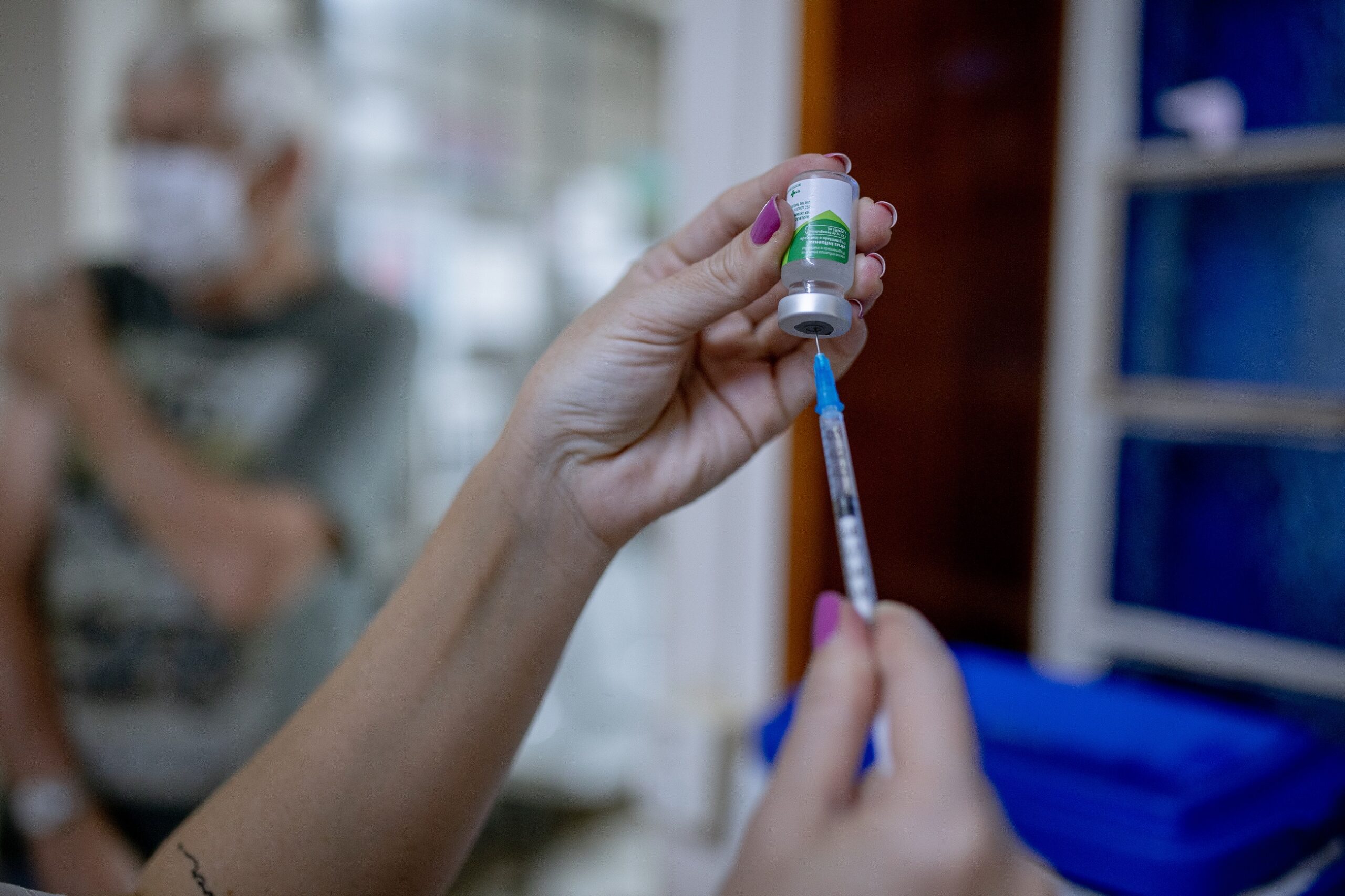 A imagem mostra uma profissional de saúde preparando uma dose de vacina. Ela está segurando um frasco de vacina com uma seringa, pronta para aspirar a vacina do frasco. No fundo, está uma pessoa parcialmente visível que parece estar esperando para receber a vacina. O ambiente parece ser uma clínica ou consultório médico. A imagem destaca a preparação e administração de vacinas, um procedimento essencial no cuidado de saúde. A imagem não permite identificar o tipo de vacina que está sendo preparada.