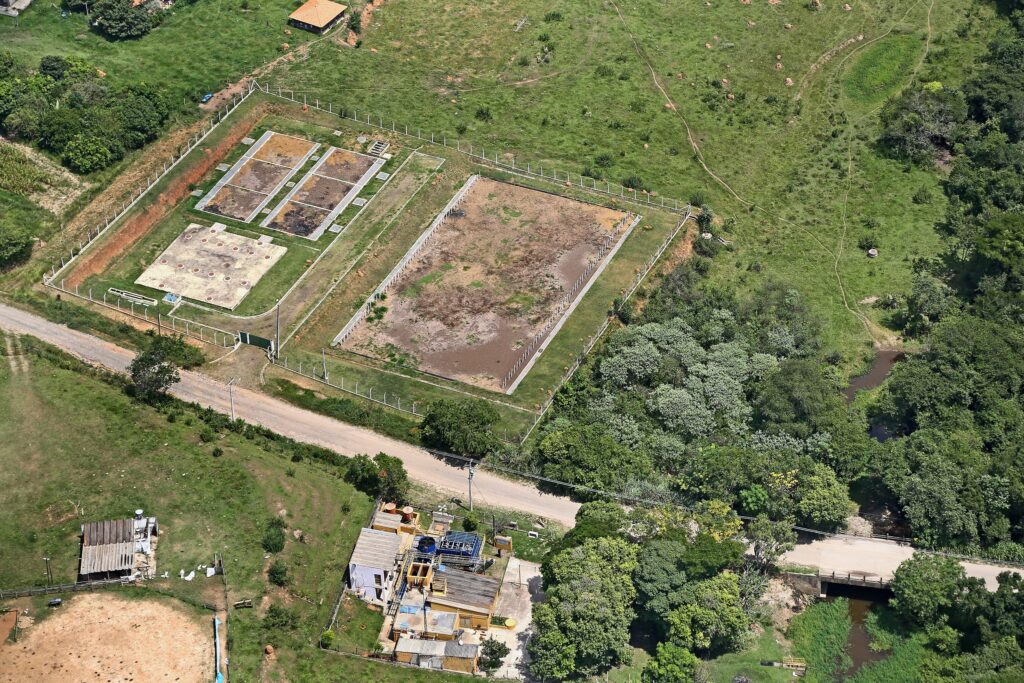 A imagem aérea mostra uma estação de tratamento de esgoto em uma área rural. A estação parece ser relativamente pequena, composta por diversos tanques de tratamento, possivelmente usando um sistema de tratamento de lagoas de estabilização. É possível observar diferentes estágios de tratamento, com tanques de tamanhos e formatos variados. A área é cercada e há uma estrada próxima. Há também outras estruturas menores próximas, que podem ser edifícios auxiliares ou instalações relacionadas à estação de tratamento. A vegetação ao redor é predominantemente verde, com áreas de pastagem e mata. Um pequeno curso d'água também é visível na parte inferior da imagem, com uma pequena ponte cruzando-o. A imagem sugere uma localização em uma região com clima tropical ou subtropical, dado o tipo de vegetação. A qualidade da imagem permite uma boa visualização da infraestrutura da estação, mas não permite uma análise detalhada dos processos de tratamento utilizados.