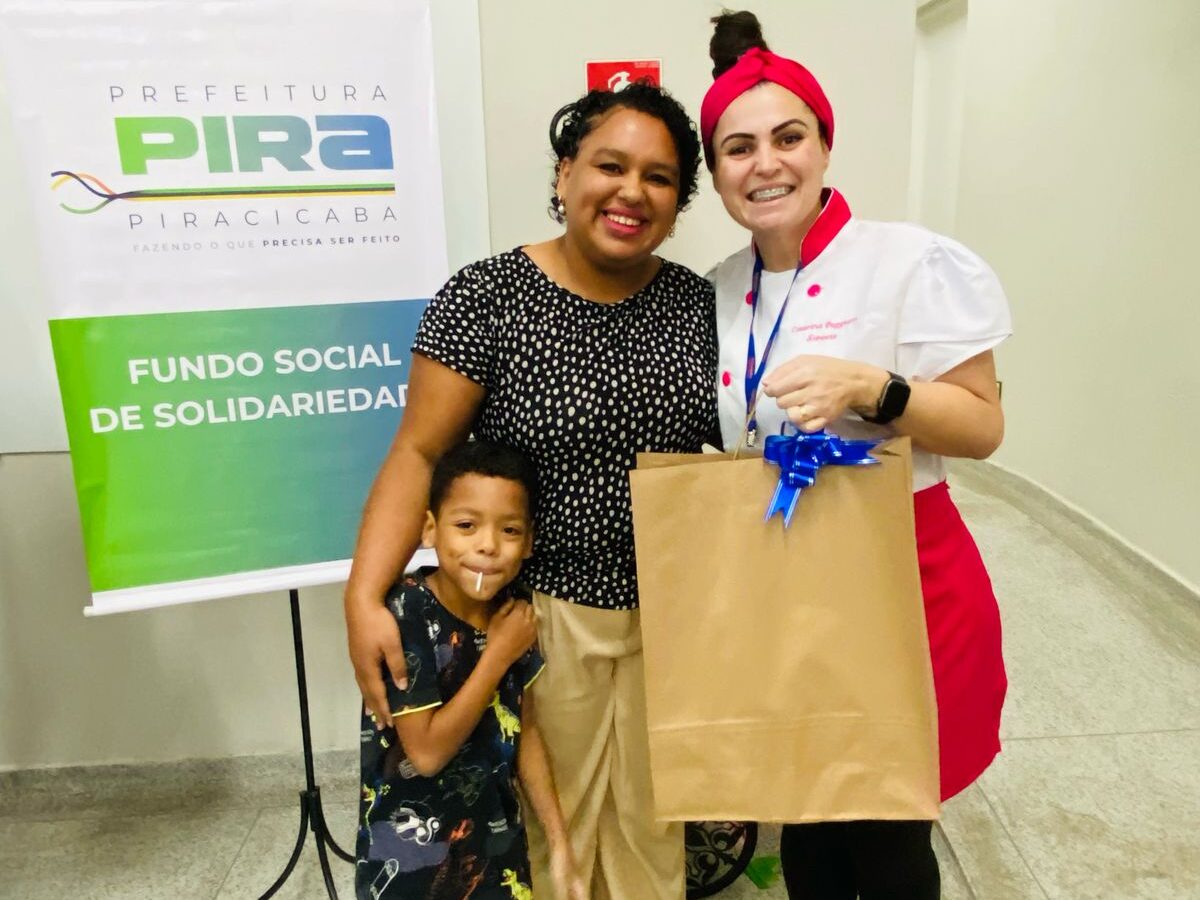 Mulher negra sorridente, ao lado de seu filho pequeno e de uma instrutora vestida com touca vermelha e uniforme de confeitaria, posam com um grande saco de papel entregue como kit de incentivo. Ao fundo, banner da Prefeitura de Piracicaba e do Fundo Social de Solidariedade.