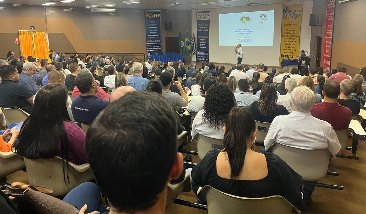 A imagem mostra uma palestra ou apresentação em um auditório. Há um grande número de pessoas sentadas em cadeiras em fileiras, presumidamente ouvindo um palestrante. Um homem está no palco, aparentemente dando uma apresentação, com um projetor exibindo uma apresentação de slides. Os slides mostram informações sobre a "Avaliação dos fatores psicossociais no trabalho", bem como informações sobre a instituição que está realizando a apresentação, possivelmente uma universidade ou faculdade, com base nas siglas e informações visíveis ("POSEEP", "EEP", "FUMEP"). Há também informações sobre os cursos oferecidos pela instituição. Em resumo, a imagem captura um evento acadêmico ou corporativo de treinamento, focado em saúde ocupacional e fatores psicossociais no trabalho. A grande quantidade de participantes sugere um evento importante e bem-sucedido.