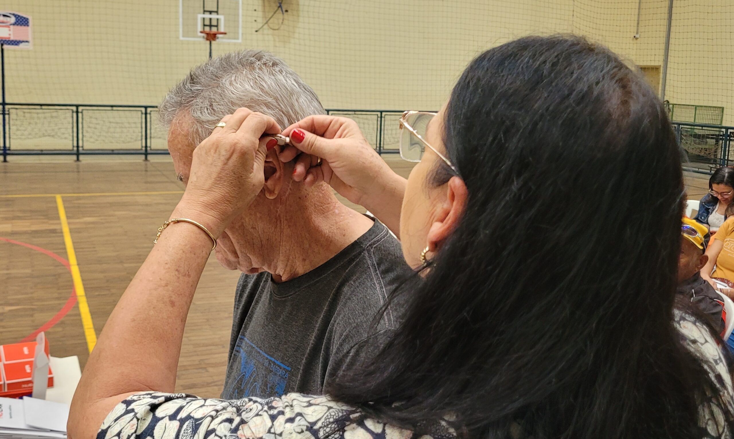 A imagem mostra uma mulher colocando um aparelho auditivo no ouvido de um homem idoso. A cena se passa em uma quadra de esportes, com uma quadra de basquete visível ao fundo. A mulher parece ser uma profissional de saúde ou alguém que está ajudando o homem a usar o aparelho. A ação é cuidadosa e atenta, sugerindo um ato de assistência. A iluminação é natural e o ambiente parece ser um local público, possivelmente um centro comunitário ou uma clínica móvel. O foco da imagem é o ato de colocar o aparelho auditivo, destacando a interação entre as duas pessoas e a assistência prestada. Não há outros detalhes relevantes na imagem além do que foi descrito.