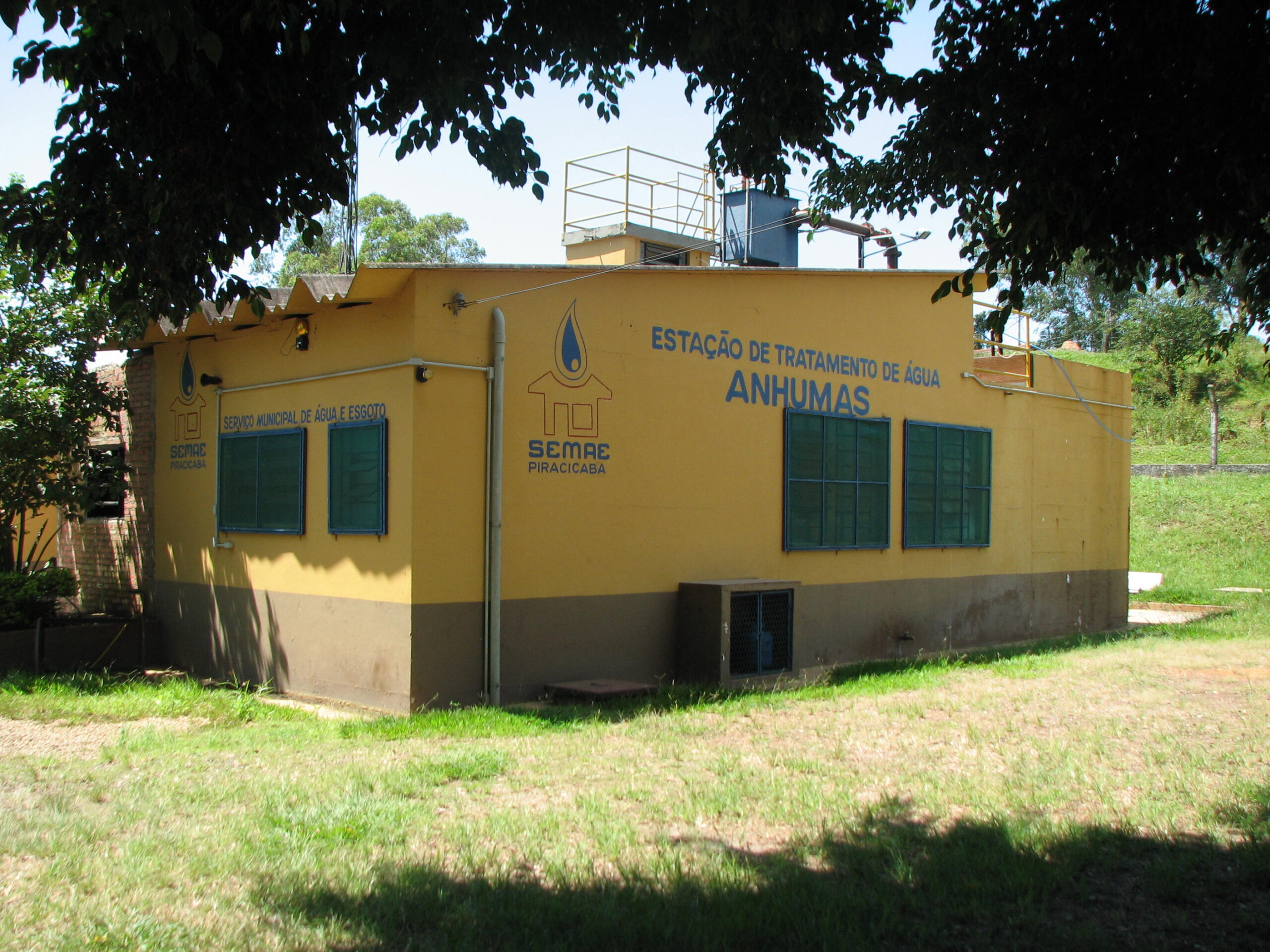 A imagem mostra uma estação de tratamento de água localizada em Anhumas. A edificação é de cor amarela com detalhes em marrom na parte inferior, e possui janelas com grades verdes escuras. Na fachada, há uma placa com o nome da estação: "Estação de Tratamento de Água Anhumas". Também é visível a logomarca e o nome da empresa responsável pela operação, "SEMAE Piracicaba", que significa Serviço Municipal de Água e Esgoto de Piracicaba. A estação parece ser relativamente pequena, sugerindo que atende a uma comunidade de tamanho moderado. A imagem também mostra uma pequena estrutura elevada na parte superior do prédio, provavelmente parte do sistema de tratamento. O ambiente ao redor é composto por grama e árvores, indicando um local provavelmente rural ou suburbano.
