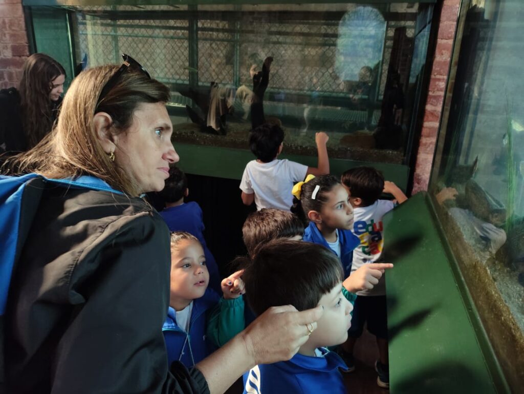 A foto mostra um grupo de crianças e adultos observando um aquário. O aquário parece estar em um ambiente educacional ou de museu, dado o ambiente e a presença de crianças com uniformes. Uma mulher com cabelos castanhos longos está no centro da imagem, ligeiramente virada para a direita. Ela está usando uma jaqueta escura e parece estar interagindo com as crianças. Ela parece ser uma professora ou guia, dada sua posição e interação com as crianças. Há várias crianças ao redor dela, algumas olhando para o aquário e outras interagindo entre si. As crianças parecem estar usando uniformes escolares, sugerindo uma excursão escolar ou uma visita guiada. O aquário é grande e contém água e algumas plantas aquáticas visíveis. Não é possível ver com clareza o que há no aquário, mas parece conter peixes ou outros animais aquáticos. Ao fundo, há outras pessoas, uma das quais parece ser outra adulta, possivelmente outra professora ou guia. A imagem é bem iluminada e mostra detalhes claros das pessoas e do ambiente. O foco está principalmente nas crianças e na mulher com cabelos compridos no centro, mas o aquário e o ambiente também são visíveis com clareza. A cena geral sugere uma atmosfera tranquila e educativa.
