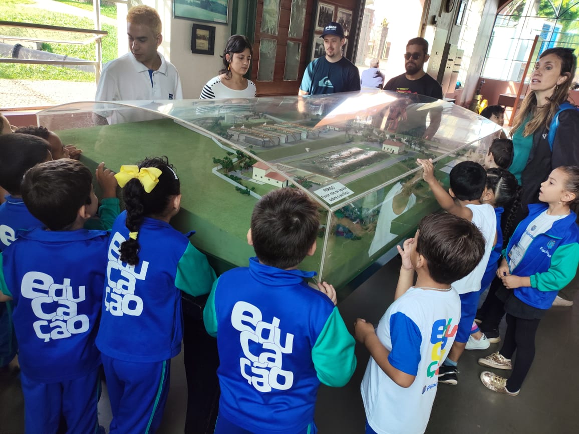 A imagem mostra uma turma de crianças em idade escolar, aparentemente em uma excursão a um museu ou centro de exposições. Elas estão reunidas em torno de um modelo em escala de uma planta industrial ou urbana, contido em uma caixa de acrílico. As crianças vestem uniformes azuis com a palavra "Educação" escrita repetidamente nas costas. Há adultos presentes, possivelmente professores ou monitores, alguns observando as crianças e outros interagindo com o modelo. O ambiente parece ser interno, com paredes de tijolos à vista e janelas que permitem a entrada de luz natural. O local parece ser limpo e organizado. A imagem sugere uma aula prática ou visita educativa, com foco no aprendizado através da observação e interação com o modelo. A atenção das crianças está voltada para o modelo, indicando engajamento com a atividade. Há pelo menos dois adultos vestidos de forma casual e outros dois com aparência mais formal, possivelmente funcionários do local. A imagem como um todo sugere uma experiência educativa positiva e envolvente para as crianças.