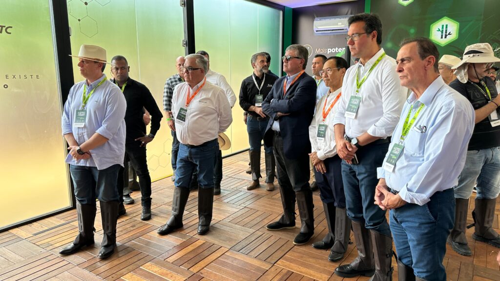A imagem mostra um grupo de pessoas reunidas em uma sala com piso de madeira e grandes janelas de vidro. Os participantes estão usando crachás e cordões, e muitos estão vestidos de maneira casual de negócios, combinando com botas de borracha. O ambiente sugere um evento profissional ou uma visita técnica, possivelmente relacionado à agricultura ou tecnologia, conforme indicado pelo texto nos crachás e pela sinalização ao fundo. Há um logotipo com as letras "CTC" e a frase "Mais poder", indicando um foco em inovação e capacidade no setor.