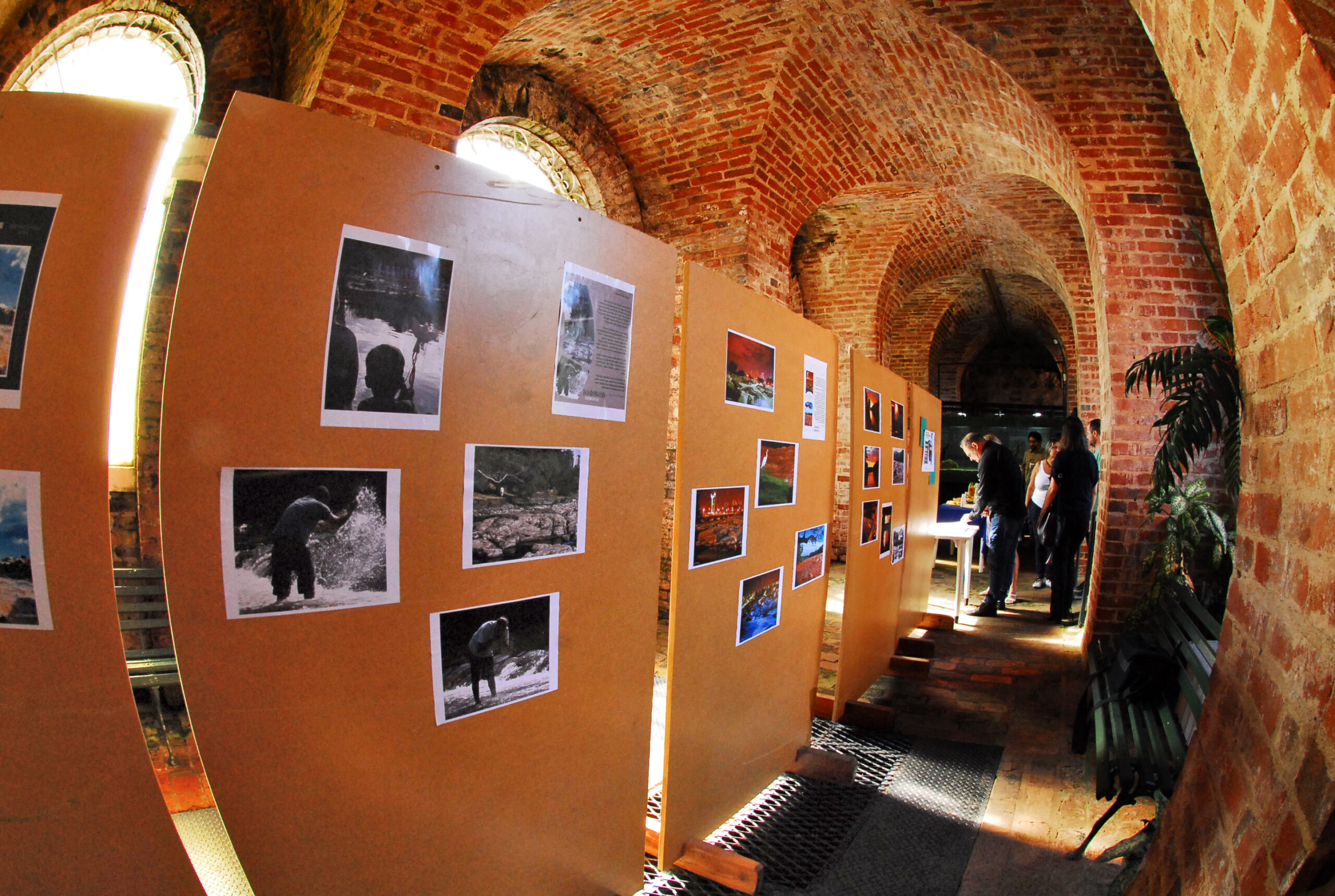 A imagem mostra uma exposição fotográfica em um local com arquitetura de tijolos aparente. A exposição está montada em painéis de cortiça ou material similar, dispostos em sequência, exibindo diversas fotografias. As fotos parecem retratar paisagens naturais, possivelmente com foco em água, e algumas imagens parecem mostrar pessoas em atividades ao ar livre. O local da exposição é um ambiente subterrâneo ou semi-subterrâneo, com arcos de tijolos que sugerem uma estrutura antiga, possivelmente um porão, uma adega ou um espaço de construção histórica. A iluminação é natural, entrando pelas janelas altas e estreitas posicionadas nos arcos de tijolo. Há também um pouco de luz artificial, visível na área onde algumas pessoas estão reunidas perto de uma mesa. Algumas pessoas podem ser vistas observando a exposição, sugerindo que o evento está acontecendo no momento da foto. Há uma atmosfera informal e tranquila. Um banco de madeira está presente em um canto, adicionando à sensação de um local informal e possivelmente histórico. A perspectiva da foto é ampla, com uma ligeira distorção de lente, provavelmente uma lente grande angular ou fisheye, que enfatiza a curvatura dos arcos e a profundidade do espaço. Em resumo, a imagem captura um momento de uma exposição fotográfica em um local histórico e incomum, com uma atmosfera tranquila e convidativa.