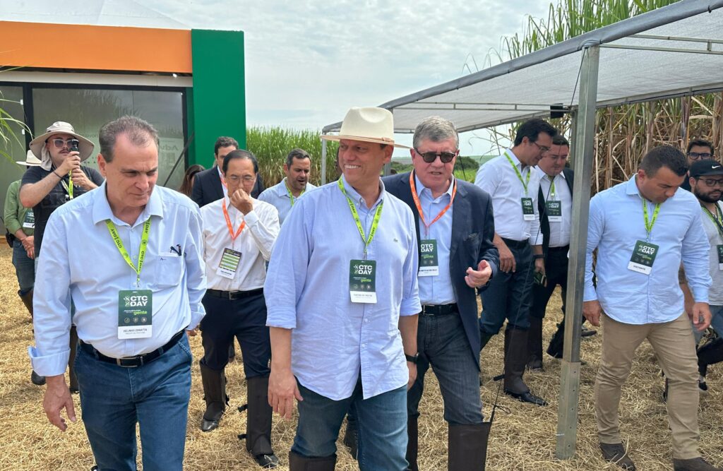 A imagem mostra um grupo de pessoas reunidas ao ar livre, possivelmente em um evento. Os participantes estão usando crachás e cordões, indicando que são convidados do encontro. Os crachás contêm os textos "CTC DAY" e "Centro de Tecnologia Canavieira". As pessoas estão vestidas de maneira casual e semi-formal, com alguns usando chapéus e botas adequadas para um ambiente externo. No fundo, há um prédio com cores verde e laranja, além de plantas de cana-de-açúcar, sugerindo que o evento pode estar relacionado à agricultura ou tecnologia no setor sucroenergético.