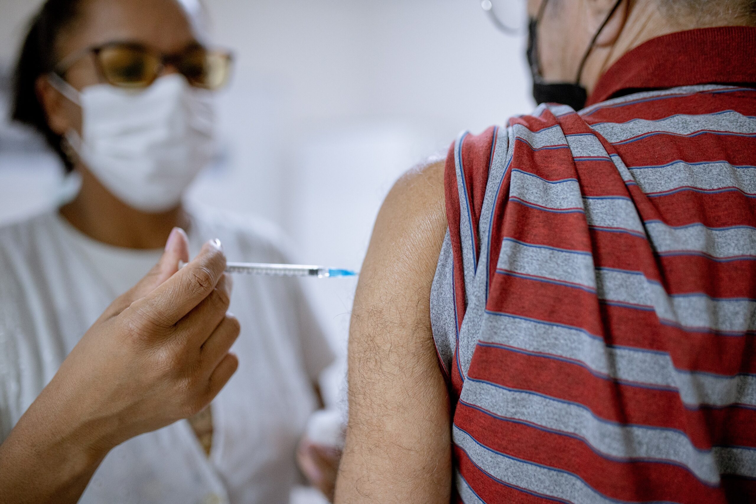A imagem mostra uma profissional de saúde aplicando uma injeção no braço de um paciente. Ambos estão usando máscaras faciais. A profissional de saúde está usando óculos de proteção e uma bata branca. O paciente está vestindo uma camisa listrada vermelha e cinza. A imagem foca no braço do paciente e na seringa. A cena sugere que a injeção é uma vacina ou algum tipo de medicamento injetável. A imagem captura um momento de um procedimento médico comum, destacando a importância da segurança e da higiene em ambientes de saúde. A máscara, os óculos de proteção e as luvas (embora não totalmente visíveis) indicam uma preocupação com a prevenção de infecções. A qualidade da imagem é boa o suficiente para observar os detalhes, mas não permite uma identificação precisa do tipo de vacina ou medicamento sendo administrado. O foco na ação da aplicação da injeção e a visibilidade parcial dos rostos dos envolvidos contribuem para um tom de privacidade e respeito à situação.