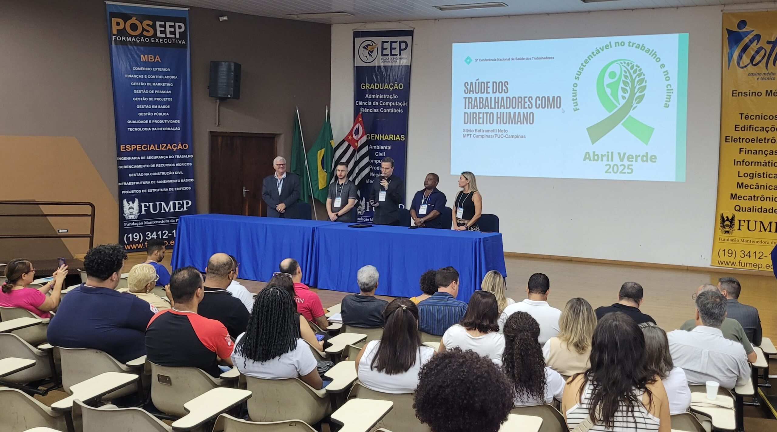 A imagem mostra uma conferência ou apresentação em um auditório. Há um painel de palestrantes na frente de uma plateia numerosa. A apresentação parece focar em "Saúde dos Trabalhadores como Direito Humano", como indicado no slide projetado na tela. O evento também parece estar conectado ao "Abril Verde 2025", uma campanha de conscientização sobre segurança e saúde no trabalho. Detalhes observáveis na imagem incluem: * *Painel de palestrantes:* Cinco pessoas estão sentadas em uma mesa, provavelmente os palestrantes do evento. * *Plateia:* Um grande número de pessoas está sentado em cadeiras, assistindo à apresentação. * *Slides de apresentação:* A tela mostra um slide com o tema da conferência e o logotipo do "Abril Verde 2025". Há também informações sobre a organização do evento e os palestrantes. * *Banner da FUMEP:* Um banner grande na lateral da sala destaca a Fundação Mantenedora da PUC-Campinas (FUMEP), indicando que a instituição pode estar patrocinando ou organizando o evento. O banner também lista cursos oferecidos pela FUMEP, incluindo MBA e especializações. * *Ambiente:* O auditório parece ser uma sala de conferências ou um auditório universitário, com cadeiras organizadas em fileiras. Em resumo, a imagem captura um momento de uma conferência sobre saúde e segurança do trabalhador, possivelmente organizada ou patrocinada pela FUMEP, no contexto da campanha "Abril Verde 2025". A imagem sugere um evento profissional e informativo.