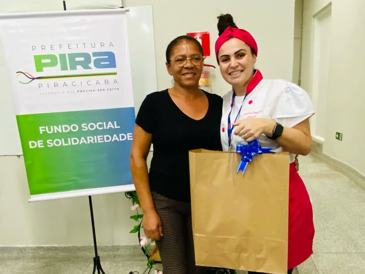 Duas mulheres sorriem em frente ao banner do Fundo Social. Uma delas, vestindo camiseta preta, recebe um saco de papel com laço azul da instrutora do curso, vestida com touca e jaleco de confeitaria.
