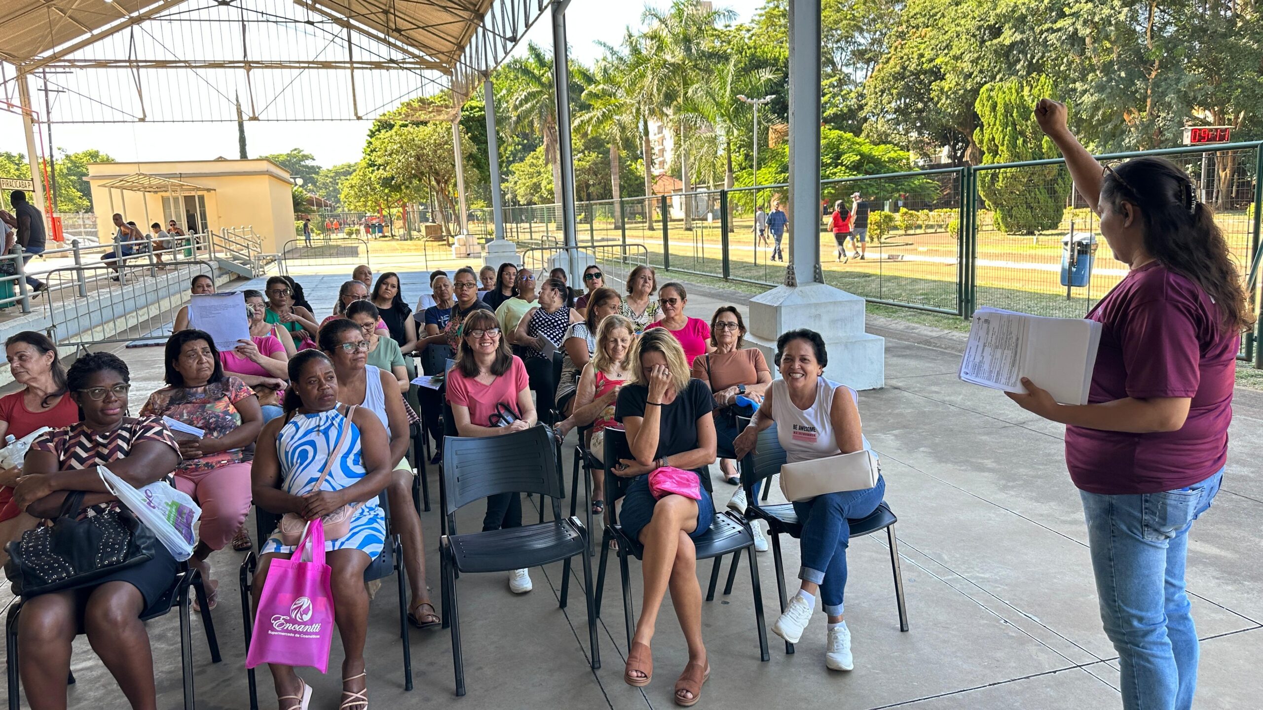 A imagem mostra um grupo de mulheres sentadas em cadeiras em um ambiente externo, sob uma estrutura coberta. Uma mulher está de pé, de costas para a câmera, segurando alguns papéis e aparentemente conduzindo uma apresentação ou reunião. As mulheres sentadas parecem estar atentas e algumas estão interagindo entre si. O local parece ser um parque ou praça pública, com árvores e grama ao fundo. As mulheres estão vestidas de forma casual, e há alguns objetos pessoais visíveis, como bolsas e sacolas. O ambiente sugere uma reunião comunitária, uma palestra, ou um evento semelhante voltado para um público feminino. A atmosfera parece calma e organizada.
