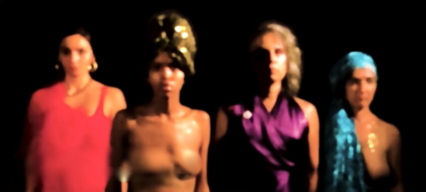 Quatro mulheres em pé, lado a lado, com roupas coloridas e iluminação baixa, aparecem levemente desfocadas contra fundo preto.