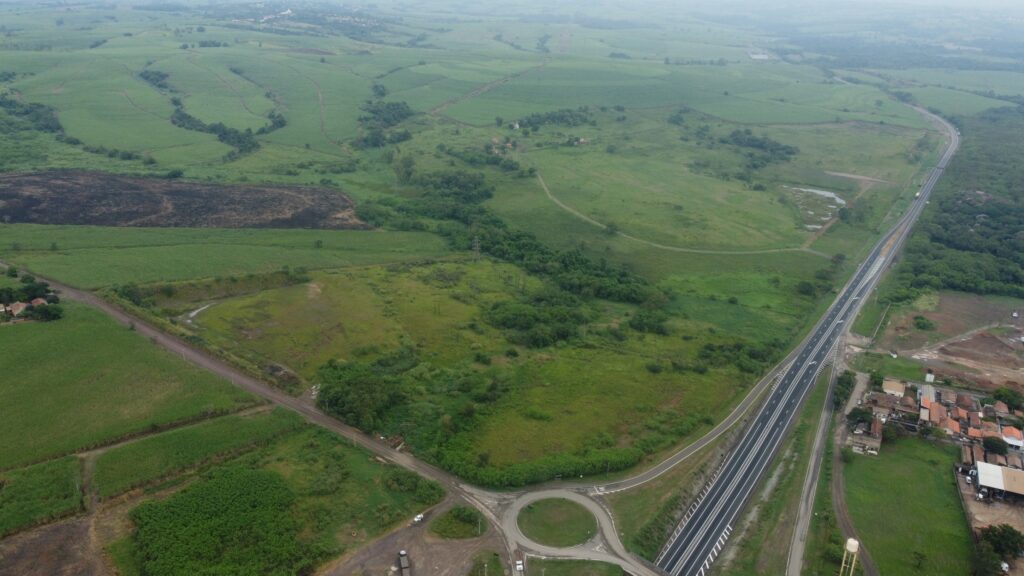 A imagem apresenta uma vista aérea de um cenário rural, composto por campos verdes, áreas arborizadas e uma rodovia que corta diagonalmente o espaço, indo do canto inferior direito ao canto superior direito. Há uma rotatória visível próxima ao centro inferior da imagem, cercada por algumas construções e casas no canto inferior direito, perto da rodovia. Os campos estão divididos em diferentes seções, algumas destinadas a agricultura. Uma área mais escura aparece na paisagem, sugerindo que pode ter sido queimada ou desmatada. O relevo é predominantemente plano, com algumas colinas suaves ao fundo, dando uma sensação de tranquilidade e harmonia entre vegetação natural e desenvolvimento de infraestrutura.