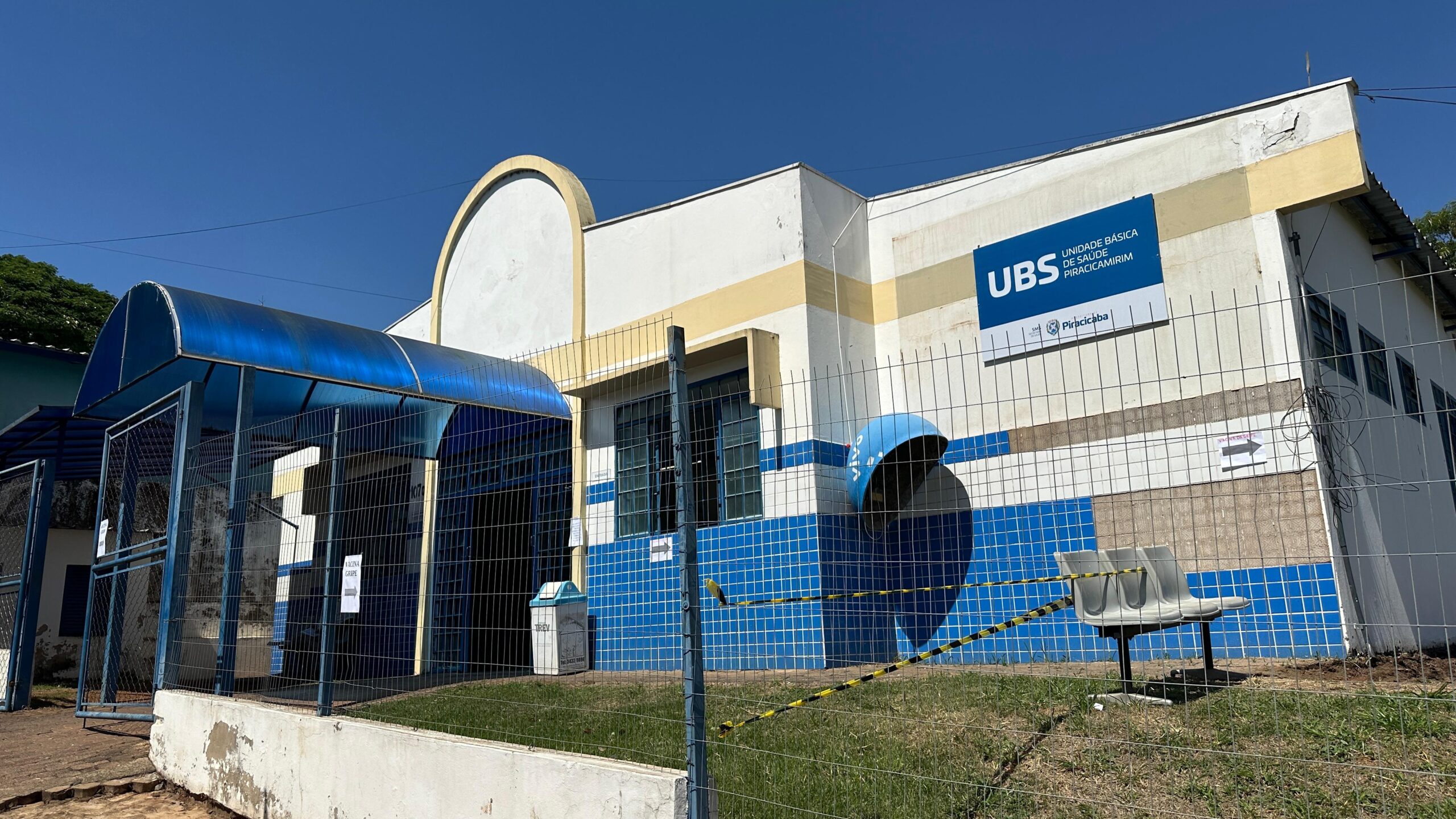 A imagem mostra uma Unidade Básica de Saúde (UBS) em Piracicaba, aparentemente fechada ou com acesso restrito. Vamos analisar os detalhes: * *Edifício:* O prédio é de estrutura simples, com arquitetura comum a unidades de saúde públicas brasileiras. Apresenta uma fachada predominantemente branca e amarela, com revestimento de azulejos azuis na parte inferior. Há um pequeno toldo azul na entrada, sugerindo um esforço para proteger os usuários do sol. O estado de conservação parece razoável, embora haja alguns sinais de desgaste visíveis. * *Segurança:* A UBS está cercada por uma cerca de arame, indicando um controle de acesso. Há também uma fita de isolamento amarela e preta na frente, reforçando a restrição de acesso. Isso sugere que a unidade pode estar temporariamente fechada, em manutenção, ou operando com restrições devido a algum motivo (pandemia, por exemplo). * *Sinalização:* Uma placa grande e visível identifica a unidade como "UBS - Unidade Básica de Saúde de Piracicaba". Há também um logotipo menor que provavelmente representa o município ou o sistema de saúde local. * *Mobiliário externo:* Dois bancos de plástico estão posicionados do lado de fora, provavelmente para uso de pacientes ou visitantes quando a unidade estiver aberta. * *Contexto:* A imagem sugere um ambiente urbano ou peri-urbano, com vegetação esparsa ao redor. Em resumo, a imagem mostra uma UBS em Piracicaba com acesso aparentemente restrito, provavelmente devido a algum motivo temporário. A imagem não fornece informações suficientes para determinar com certeza o motivo da restrição de acesso.