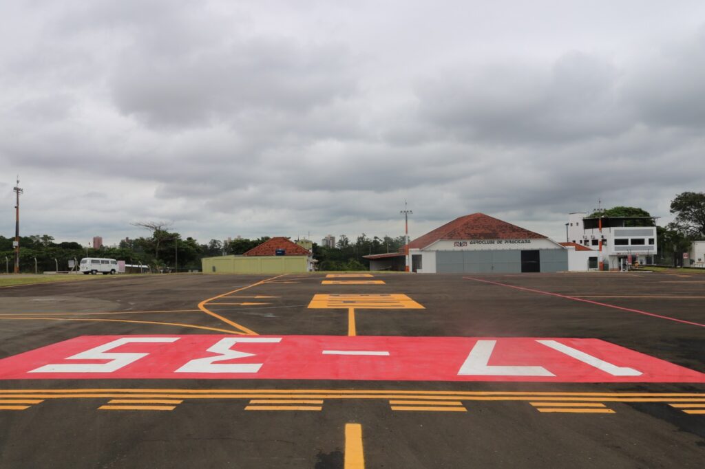 A imagem mostra um aeródromo com uma pista de pouso marcada com os números "17-35" em texto branco destacado sobre um fundo vermelho. A pista também possui marcações amarelas e laranjas, indicando áreas específicas para operações. Ao fundo, há edifícios, incluindo um com o texto "AEROCLUBE DE PIRACICABA" em destaque. Também é possível ver uma van branca estacionada no lado esquerdo da imagem. O céu está nublado, criando um ambiente levemente cinzento.