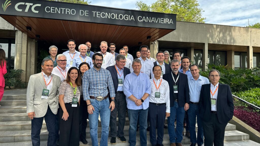 A imagem mostra um grupo de pessoas reunidas na escadaria em frente a um prédio com uma placa que diz "CTC Centro de Tecnologia Canavieira". Os participantes estão usando crachás e cordões, indicando que estão participando de um evento ou conferência no centro tecnológico. O prédio está rodeado de vegetação, e as pessoas estão vestidas de maneira casual de negócios.