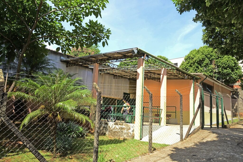 Fotografia colorida da entrada do Centro Cultural Izaíra Aparecida Barbosa, também conhecido como Zazá. O prédio é térreo, com cobertura metálica e colunas em tons claros. Na frente, há uma pequena área com plantas e uma árvore sombreando parte da calçada. A fachada é protegida por um portão e cercado de arame. Duas pessoas conversam na entrada, ao lado de uma moto.