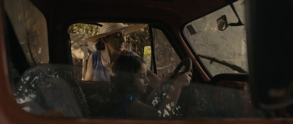 Mulher com chapéu de palha observa uma menina ao volante de uma caminhonete antiga, vista pela janela do veículo.