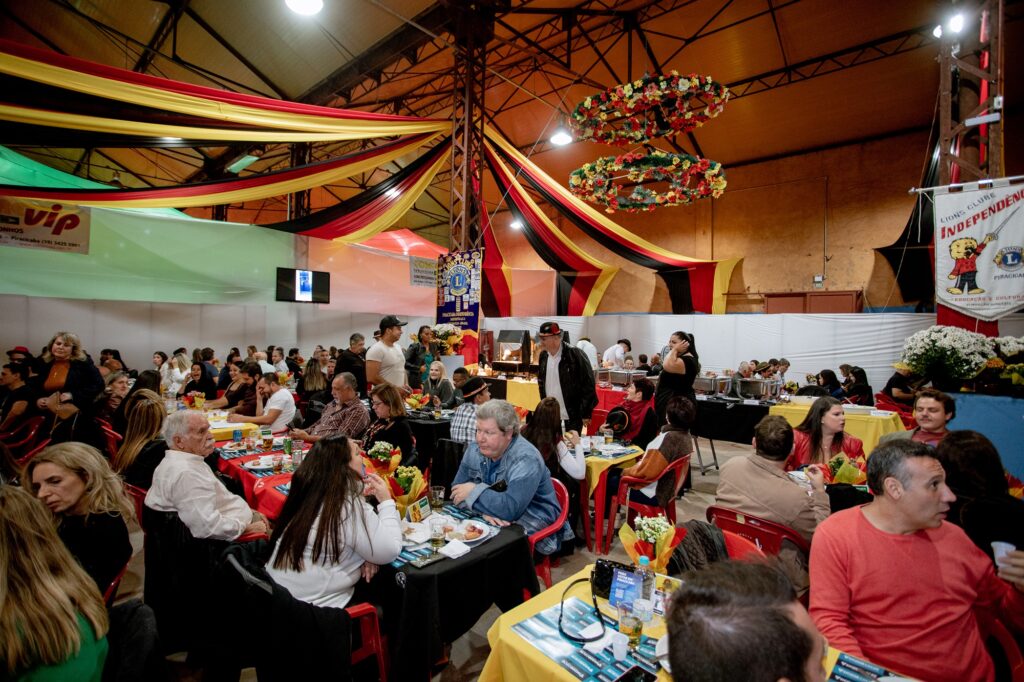 A imagem mostra um grande evento em ambiente interno, com muitas pessoas sentadas em mesas, aparentemente participando de um banquete ou celebração. As mesas estão decoradas com toalhas coloridas e centros de mesa florais. O teto é adornado com tecidos drapeados em vermelho, amarelo e preto, além de decorações suspensas feitas de flores. Há banners nas paredes, incluindo um que diz "Lions Clube Independência" e outro com a palavra "VIP." Também é possível ver uma área de buffet onde as pessoas estão se servindo.