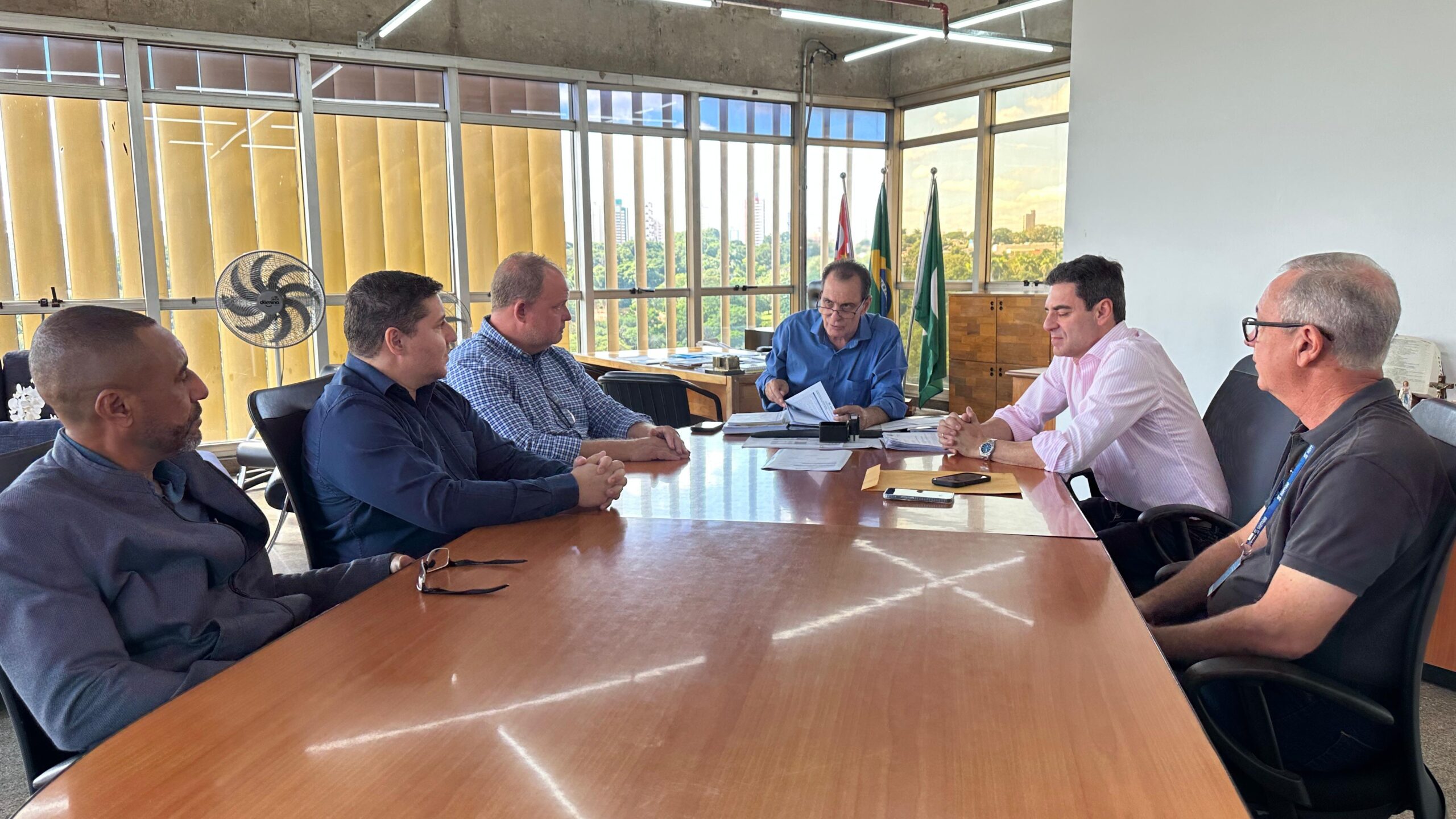 A imagem mostra uma reunião em uma sala de conferências. Seis homens estão sentados ao redor de uma mesa de madeira retangular. A sala tem grandes janelas que mostram uma vista arborizada com prédios ao fundo. Duas bandeiras, possivelmente brasileira e de um estado ou município, estão penduradas atrás dos participantes. Os homens parecem estar envolvidos em uma discussão séria ou em uma reunião de negócios, com papéis e documentos espalhados na mesa. Um ventilador de teto está visível no canto esquerdo da imagem. A atmosfera parece formal e profissional. Não há mais informações disponíveis na imagem para fornecer detalhes adicionais sobre os participantes ou o propósito da reunião.
