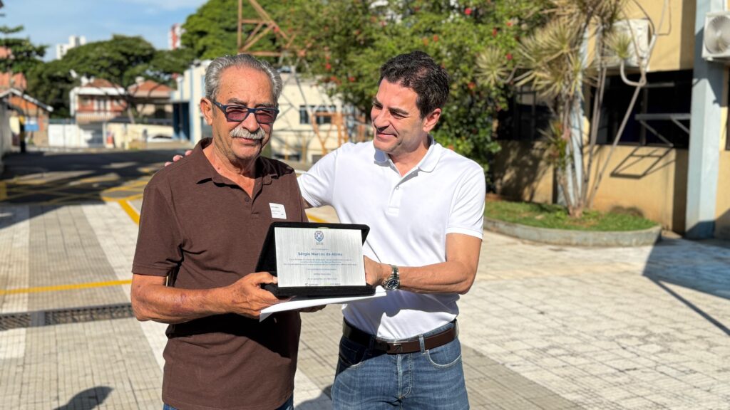 A imagem mostra dois homens, um mais velho e um mais novo, em um ambiente externo. O homem mais velho, com óculos e camisa marrom, está recebendo uma placa de homenagem das mãos do homem mais novo, que veste uma camisa branca e jeans. A placa contém o nome "Sérgio Marcos de Abreu" e um texto de reconhecimento. O fundo mostra uma edificação e vegetação, sugerindo um ambiente corporativo ou institucional. A cena parece ser um evento de premiação ou reconhecimento profissional, onde o homem mais novo está entregando a homenagem ao homem mais velho por seus serviços prestados. A expressão facial de ambos indica respeito e satisfação. A iluminação e a qualidade da imagem sugerem que a foto foi tirada em um dia ensolarado, com boa qualidade de câmera.