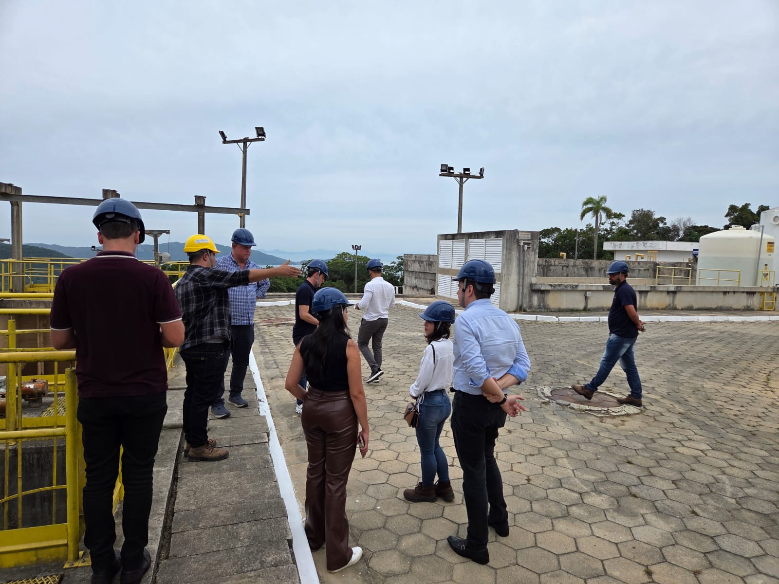 A imagem mostra um grupo de pessoas em um passeio ou visita guiada em uma instalação industrial, possivelmente uma estação de tratamento de água ou uma planta de processamento. A localização parece ser externa, em uma área pavimentada com blocos de concreto em formato hexagonal. Ao fundo, é visível uma linha de horizonte com montanhas e vegetação. O céu está nublado, sugerindo um dia sem sol intenso. *Detalhes sobre as pessoas:* * O grupo é composto por aproximadamente dez indivíduos, todos vestindo capacetes de segurança, indicando um ambiente de trabalho ou visita técnica. * A maioria dos capacetes são azuis escuros, exceto dois que são amarelos. * As vestimentas são informais, mas apropriadas para um ambiente industrial. Há uma mistura de calças jeans, calças sociais e camisas polo. Uma mulher usa calças de couro marrom. * Dois homens parecem estar conduzindo a visita, um deles usando um capacete amarelo e apontando para algo no horizonte. * As pessoas estão distribuídas em pequenos grupos, sugerindo conversas e explicações durante a visita. * Um homem está caminhando sozinho um pouco distante do grupo principal. *Detalhes sobre o ambiente:* * A área é uma plataforma aberta, com uma estrutura de concreto ao redor. * Há tanques grandes e equipamentos industriais visíveis ao fundo. * Há postes de iluminação em diferentes pontos da área. * A vegetação ao fundo sugere que a instalação está localizada em um ambiente mais natural ou suburbano. * As grades amarelas sugerem proteção de segurança em torno de equipamentos ou aberturas. *Conclusão:* A imagem retrata um momento de uma visita técnica ou passeio em uma instalação industrial, onde um grupo de pessoas está recebendo informações de guias. O ambiente é seguro, com todos usando os equipamentos de proteção necessários. A atmosfera parece ser de aprendizado e observação. A localização sugere um ambiente industrial próximo à natureza.