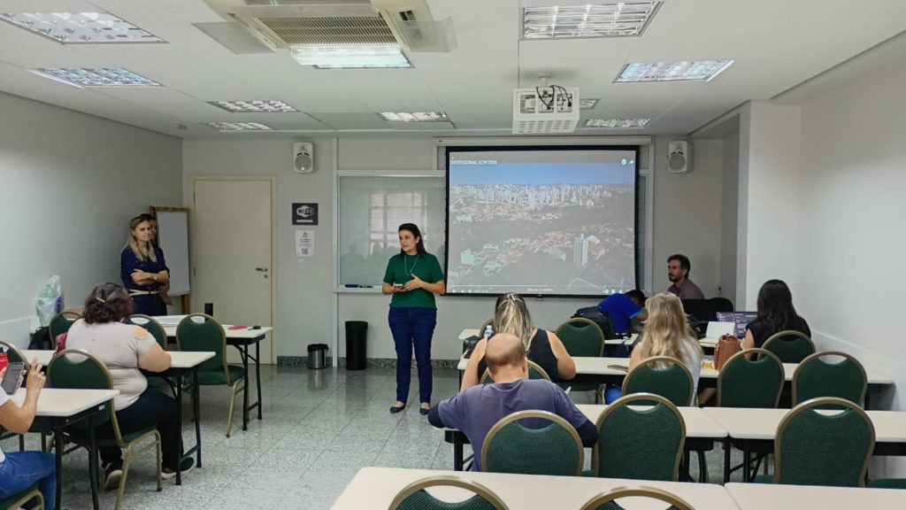 A imagem mostra uma sala de aula com várias pessoas sentadas em mesas e cadeiras. Há uma tela de projeção na frente da sala, mostrando uma imagem aérea de uma cidade. Uma mulher está de pé na frente da tela, parecendo dar uma apresentação. Outra mulher está de pé ao lado dela, possivelmente assistindo ou ajudando. Há um homem sentado perto da tela, possivelmente um assistente técnico. Os alunos estão sentados em mesas. A sala parece limpa e organizada, com iluminação adequada. O ambiente parece ser formal e acadêmico. A imagem sugere uma apresentação ou aula em andamento.