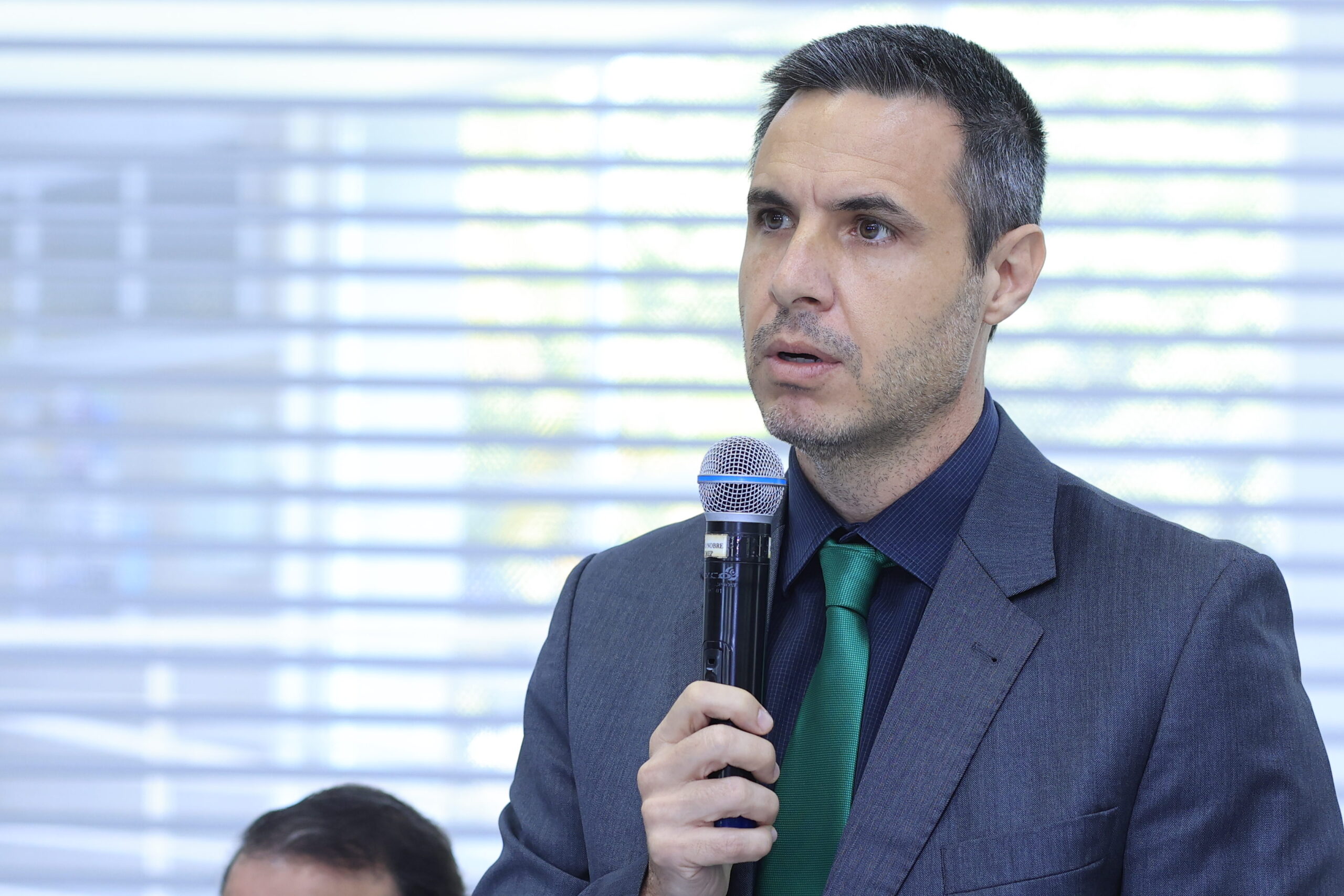 A imagem mostra um homem de meia-idade, vestindo um terno escuro e gravata verde, falando em um microfone. Ele parece estar em um ambiente profissional, possivelmente em uma conferência ou apresentação, considerando o fundo desfocado que sugere uma janela com persianas. A expressão facial dele sugere concentração e seriedade, enquanto ele fala. A qualidade da imagem é boa, com detalhes nítidos do rosto e do traje do homem. Há uma parte da cabeça de outra pessoa visível no canto inferior esquerdo, indicando que pode haver mais pessoas presentes no local. A iluminação é suave e uniforme, sem sombras fortes. Em resumo, a imagem transmite uma sensação de profissionalismo e formalidade.