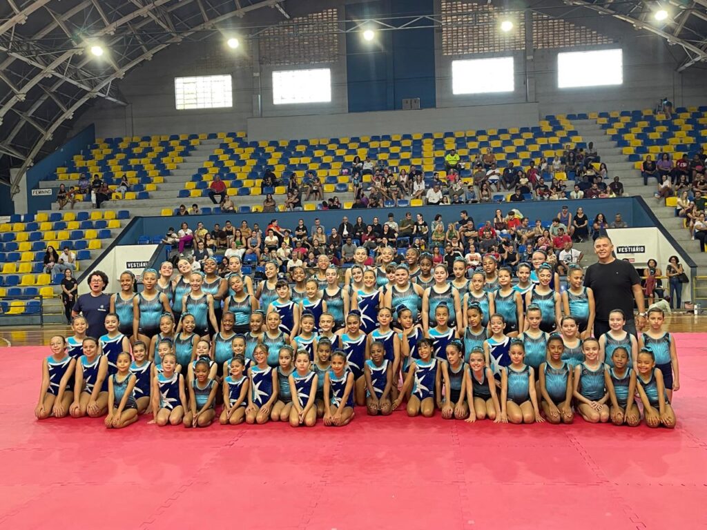 Em quadra coberta com arquibancadas, cerca de 80 meninas, todas com collants de ginástica nas cores azul e preto, posam organizadas em fileiras sobre um grande tapete vermelho. Elas participam do 1º Festival de Ginástica Artística e Acrobática. À esquerda e à direita, dois professores acompanham o grupo. Ao fundo, o ginásio Waldemar Blatkauskas está parcialmente ocupado por familiares e espectadores nas arquibancadas com assentos azuis e amarelos. A iluminação é forte e o ambiente transmite clima de alegria e celebração.