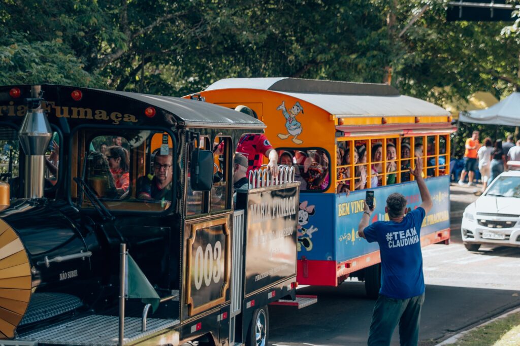 A imagem mostra um trem turístico colorido com dois vagões, um preto e um amarelo, cheio de passageiros. O vagão preto tem o número "003" e o nome "Maria Fumaça" escrito nele. O vagão amarelo tem um personagem de desenho animado pintado na lateral e o texto "BEM VINDO".