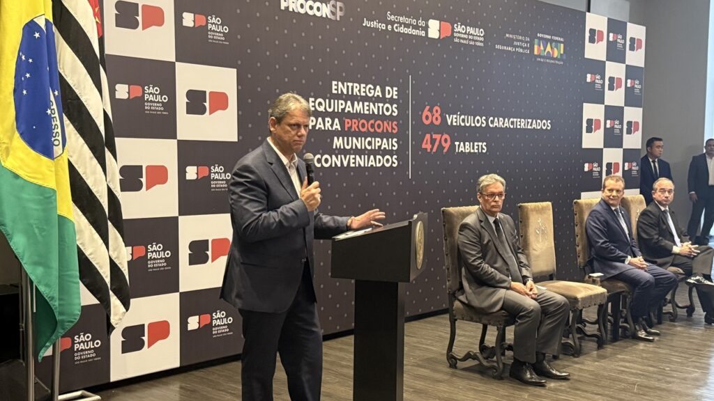 A imagem mostra uma cerimônia de entrega de equipamentos para Procons municipais conveniados no estado de São Paulo.  Um homem de terno está falando ao microfone, em frente a um painel com os logos do governo do estado e as quantidades de veículos e tablets distribuídos (68 veículos e 479 tablets). Há outras pessoas sentadas em cadeiras na frente do painel, provavelmente autoridades.  Bandeiras do Brasil e do estado de São Paulo estão visíveis ao lado do painel. O ambiente parece ser um salão de eventos ou uma sala de conferências formal.