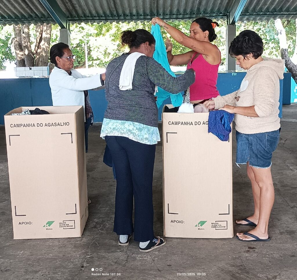 A imagem mostra quatro mulheres em uma campanha de doação de roupas. Elas estão em um espaço coberto, organizando roupas em duas grandes caixas de papelão. As caixas estão rotuladas como "Campanha do Agasalho" e indicam o apoio da Klabin. As mulheres parecem estar separando e doando as roupas. O clima parece ser ensolarado, e o ambiente é simples, ao ar livre.  A foto foi tirada em 23/05/2025 às 09:35.