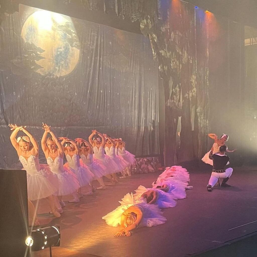 Bailarinas de vestido branco se apresentam em cena ao lado de um bailarino. Algumas estão em pé, outras ajoelhadas com braços erguidos e parte do grupo deitada em fila no chão, compondo uma coreografia visual sob luz suave.