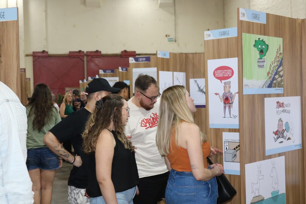 Foto colorida mostra um grupo de visitantes observando painéis de madeira vertical com charges e cartuns expostos em folhas brancas. As pessoas estão próximas dos painéis, olhando atentamente os desenhos. A maioria veste roupas casuais e há uma predominância de jovens. O espaço é fechado, com paredes altas, teto claro e iluminação artificial.