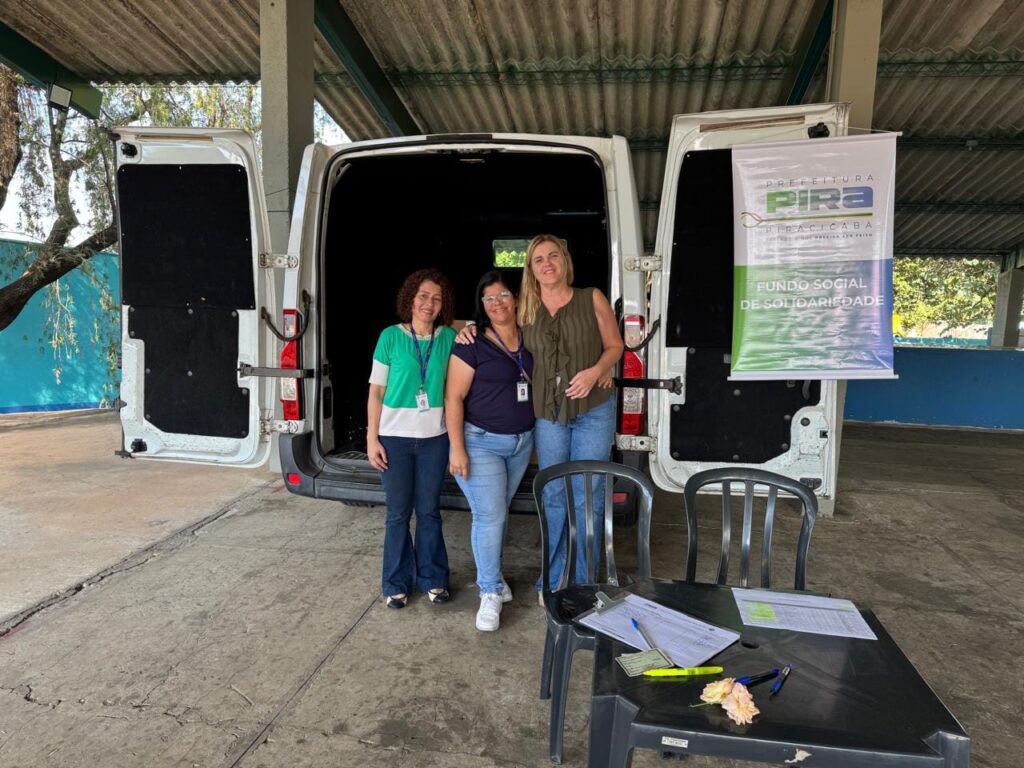 A imagem mostra três mulheres sorrindo, em pé na frente de uma van branca. Elas estão abraçadas e parecem felizes. A van tem uma placa que diz "Prefeitura Piracicaba - Fundo Social de Solidariedade". Há uma mesa pequena com alguns papéis e canetas na frente delas. O cenário parece ser um estacionamento ou área externa. O ambiente parece ser calmo e organizado. As mulheres parecem estar trabalhando juntas em algum projeto social. A imagem transmite uma sensação de comunidade e colaboração.