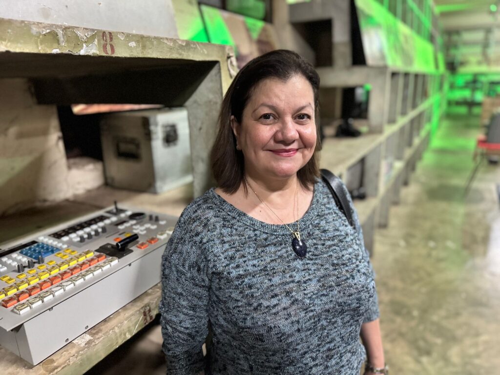 A imagem mostra uma pessoa no Museu da Imagem e do Som de Piracicaba, um espaço dedicado à preservação e exposição de elementos visuais e sonoros. Ela está vestindo um suéter azul-acinzentado e um colar com um pingente escuro. À esquerda, há um painel de controle com vários botões e interruptores coloridos—amarelos, vermelhos e azuis—além de deslizadores e botões giratórios. O fundo contém prateleiras ou compartimentos de concreto, alguns dos quais abrigam caixas metálicas ou equipamentos. A iluminação verde dá um toque marcante ao ambiente, contribuindo para a atmosfera imersiva do museu, que parece explorar a tecnologia e a história da imagem e do som.