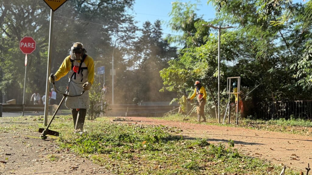 A imagem mostra dois trabalhadores realizando serviços de paisagismo ou manutenção em uma calçada. Ambos estão utilizando roçadeiras para cortar grama e ervas daninhas. O trabalhador em primeiro plano está usando equipamentos de proteção, incluindo capacete, luvas e uma viseira, e está ativamente aparando a grama. O trabalhador ao fundo também está envolvido na mesma atividade. No fundo, há uma placa com a palavra "PARE", indicando que o local pode estar em uma região de língua portuguesa. A cena acontece em uma área verde com árvores e vegetação, e há um pouco de poeira no ar devido ao trabalho de corte.