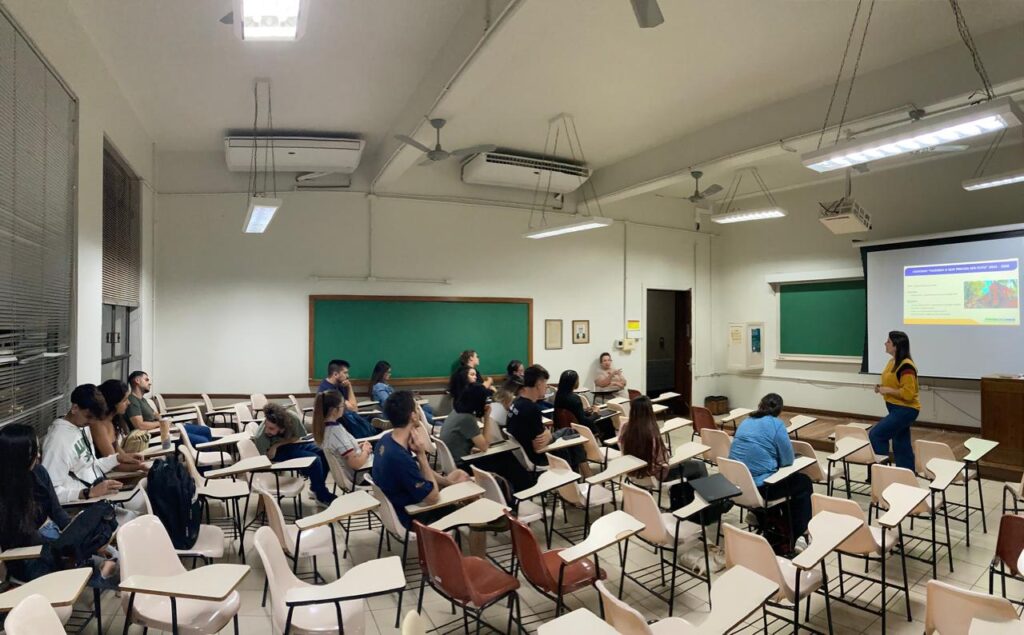 A imagem mostra uma sala de aula universitária durante uma apresentação. A sala está organizada com carteiras individuais dispostas em fileiras, e a maioria delas está ocupada por estudantes.  Há uma professora ou palestrante em pé perto de um projetor, apresentando algo em uma tela.  A tela exibe um slide com texto e imagens, sugerindo uma apresentação formal.A sala é bem iluminada, com lâmpadas fluorescentes no teto e janelas laterais com persianas. Há também aparelhos de ar condicionado instalados no teto. A decoração é simples e funcional, típica de uma sala de aula.  As paredes são claras e há um quadro-negro na parte frontal da sala.  Os estudantes parecem engajados, alguns participando ativamente, outros prestando atenção. A atmosfera é calma e focada no aprendizado.  A imagem captura um momento de uma aula ou palestra, provavelmente em uma universidade ou instituição de ensino superior.