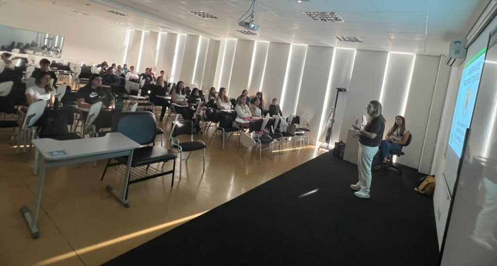 A imagem mostra uma sala de aula universitária durante uma apresentação. Há uma palestrante em pé na frente de uma tela de projeção, que exibe um slide com o que parece ser um título sobre insetos ou pragas. Ela está vestindo roupas casuais e parece estar interagindo com a audiência. Há uma outra pessoa sentada perto da palestrante, possivelmente uma assistente ou colega. A sala de aula está razoavelmente cheia de estudantes sentados em carteiras individuais dispostas em fileiras. Os alunos parecem estar atentos à apresentação. A iluminação da sala é suave e a atmosfera parece calma e focada na apresentação. A sala é moderna e bem iluminada, com paredes brancas e um teto com iluminação embutida. Há um projetor pendurado no teto. A disposição das carteiras sugere uma aula tradicional. Em resumo, a imagem captura um momento típico de uma aula ou palestra em uma universidade, com foco na apresentação e na interação entre a palestrante e os alunos. A presença da tela de projeção e o conteúdo do slide indicam uma apresentação formal com material visual.
