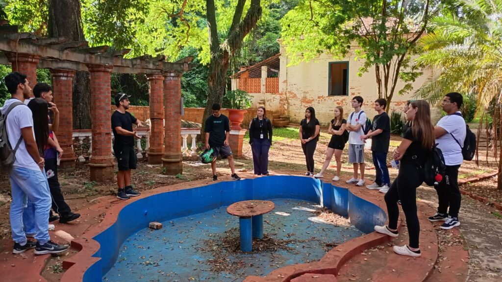 A imagem mostra um grupo de estudantes, aparentemente em uma excursão ou aula ao ar livre, reunidos em torno de uma fonte seca em um ambiente que parece ser um jardim ou parque histórico.*Detalhes da cena:** *Os estudantes:* Um grupo de cerca de 12 a 15 estudantes, com idades que parecem estar entre 15 e 20 anos, estão reunidos. Eles estão vestidos com roupas casuais. Alguns estão mais atentos a um instrutor, enquanto outros parecem mais relaxados ou conversando entre si. Há uma diversidade de gêneros e etnias no grupo.* *O instrutor:* Uma pessoa, possivelmente um professor ou guia, está no centro da atenção, parecendo explicar algo ao grupo. Ele está vestido com uma camiseta escura e shorts, e parece interagir com os estudantes de forma informal.* *O local:* O local parece ser um jardim ou parque com uma arquitetura antiga. Há uma fonte seca no centro da imagem, com uma estrutura de pedra e concreto desgastada. Uma estrutura de pergolados de tijolos vermelhos em ruínas, parcialmente destruída, está ao fundo, sugerindo um local histórico ou abandonado. Há uma casa antiga de estilo colonial à distância, também em estado de degradação. A vegetação ao redor é exuberante e tropical, com árvores grandes e folhagens densas.* *A atmosfera:* A atmosfera parece ser calma e educativa. O grupo parece engajado na atividade, sugerindo uma aula ou tour guiado. A luz natural indica que a imagem foi provavelmente tirada durante o dia.Em resumo, a imagem captura um momento de aprendizado ao ar livre, onde um grupo de estudantes está envolvido em uma atividade educativa em um ambiente histórico e natural. A imagem transmite uma sensação de história, aprendizado e interação entre os estudantes e o instrutor.