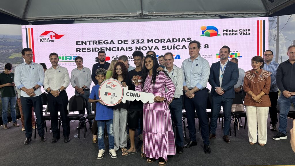 A imagem mostra uma cerimônia oficial de entrega de 332 moradias do Residencial Nova Suíça 8, em Piracicaba. Ao centro, uma família posa sorridente com uma chave simbólica da CDHU. Ao fundo, autoridades estaduais e municipais acompanham o momento. O evento integra os programas Minha Casa Minha Vida e Casa Paulista.